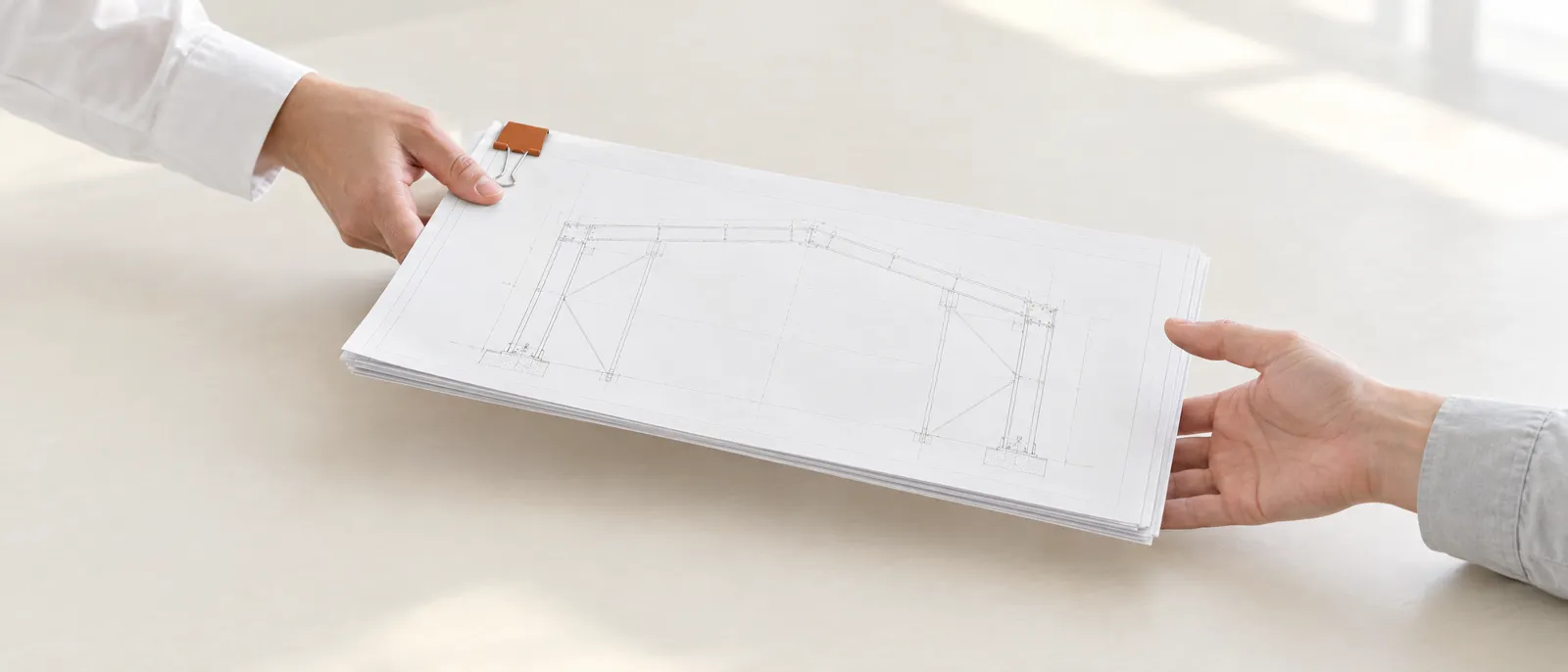 Illustration of one hand passing a squared set of portal frame drawings, held by a rust-coloured binder clip, across a pale table to another hand.